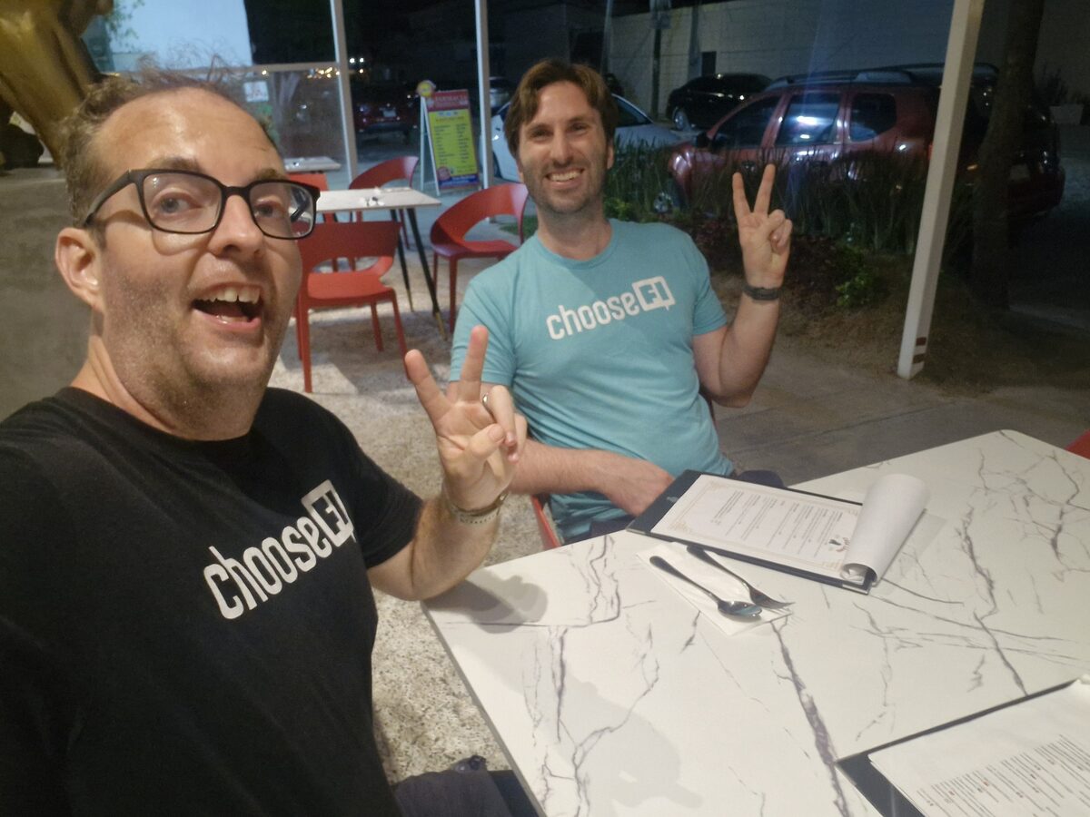 Justin and Daniel at an outdoor restaurant table at night, both in matching ChooseFI t-shirts, grinning and throwing peace signs at the camera with unopened menus on the table in front of them.