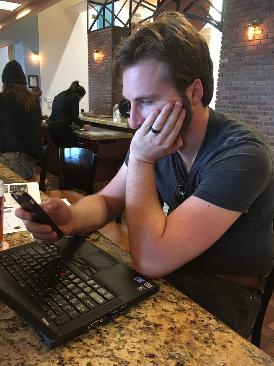 Daniel sitting at a restaurant bar counter with an open ThinkPad laptop in front of him and a phone in one hand, his chin resting in the other, looking exhausted. Other diners are visible behind him.