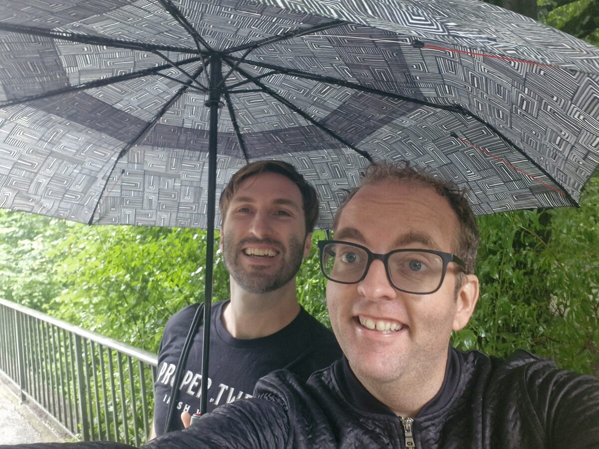 Daniel and Justin crowded under one patterned umbrella in a downpour in Germany, both grinning at the camera, wet green foliage and a railing behind them.