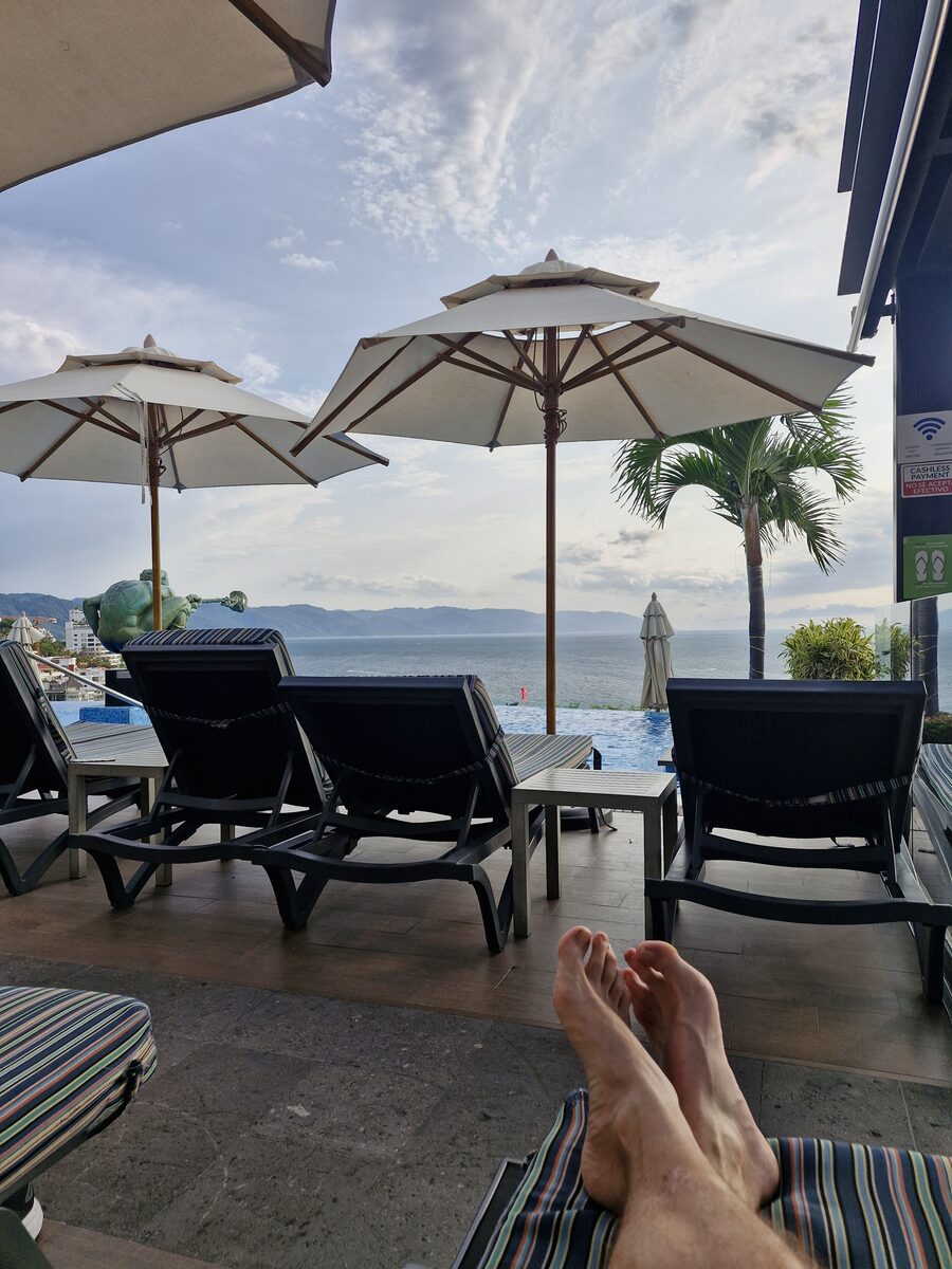 View from a poolside lounger, my bare feet propped up in the foreground, looking past empty loungers and white patio umbrellas to an infinity pool, a palm tree, and the ocean with mountains on the horizon.